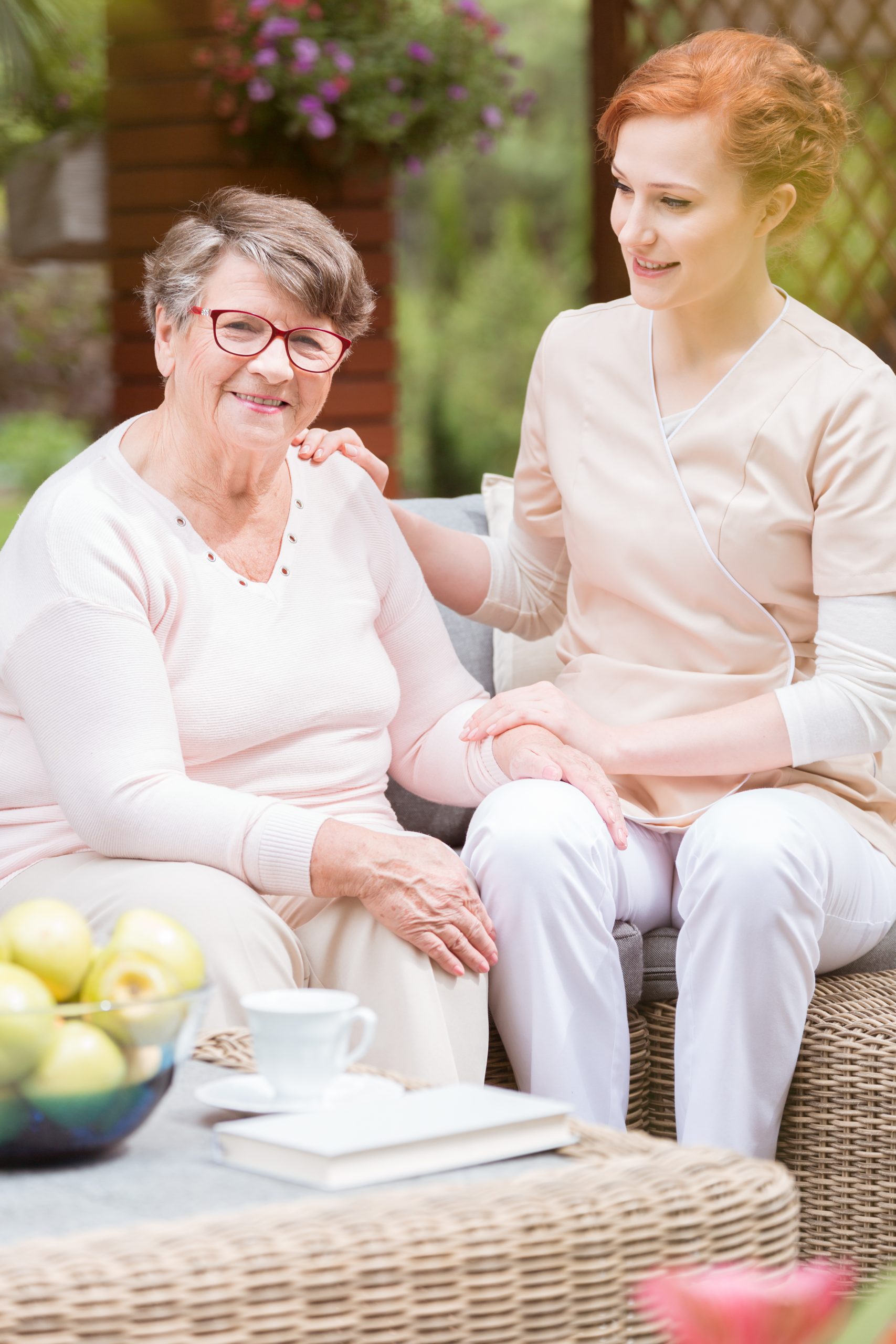 Tender professional caregiver in uniform putting her hand on a shoulder of an elderly woman during snack time on a patio of a senior home. Blurred background.